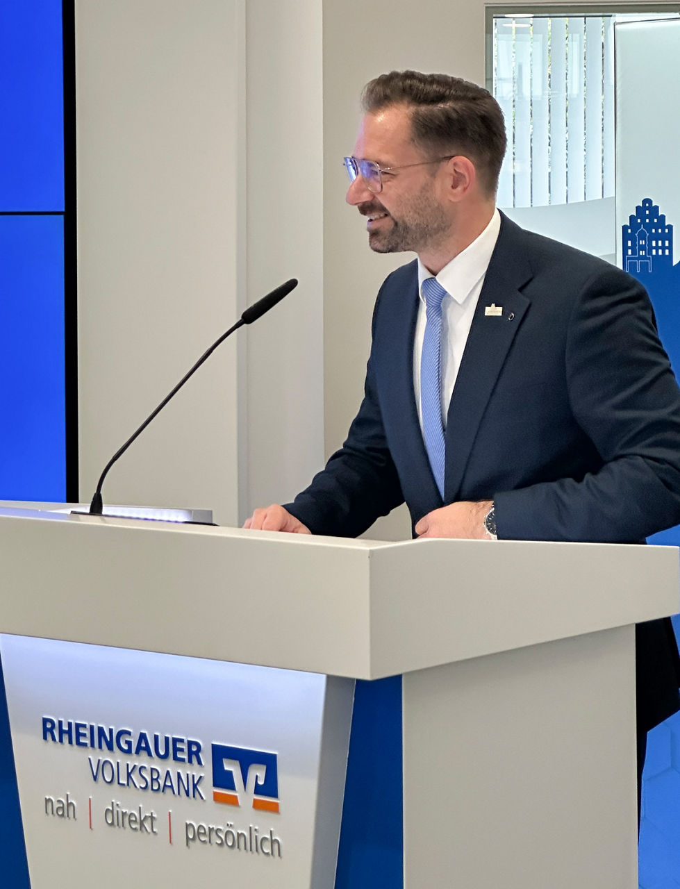 Mann im dunklen Anzug mit hellblauer Krawatte steht an einem Rednerpult mit dem Logo der Rheingauer Volksbank.