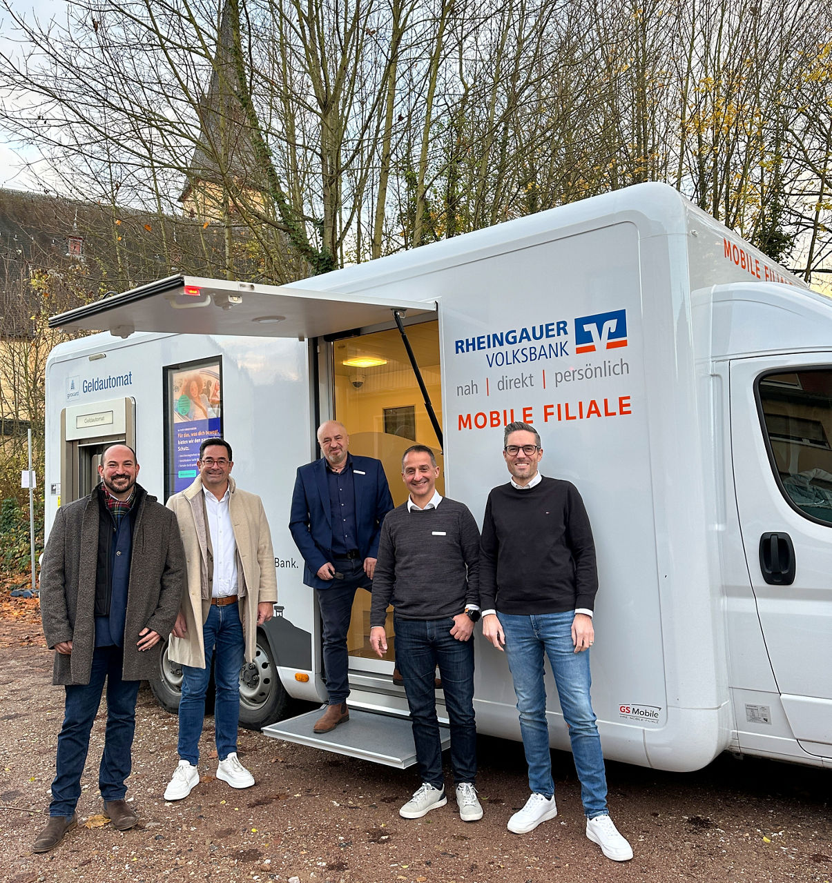 Fünf Männer stehen vor einem weißen mobilen Bankfahrzeug der Rheingauer Volksbank mit geöffnetem Seiteneingang.