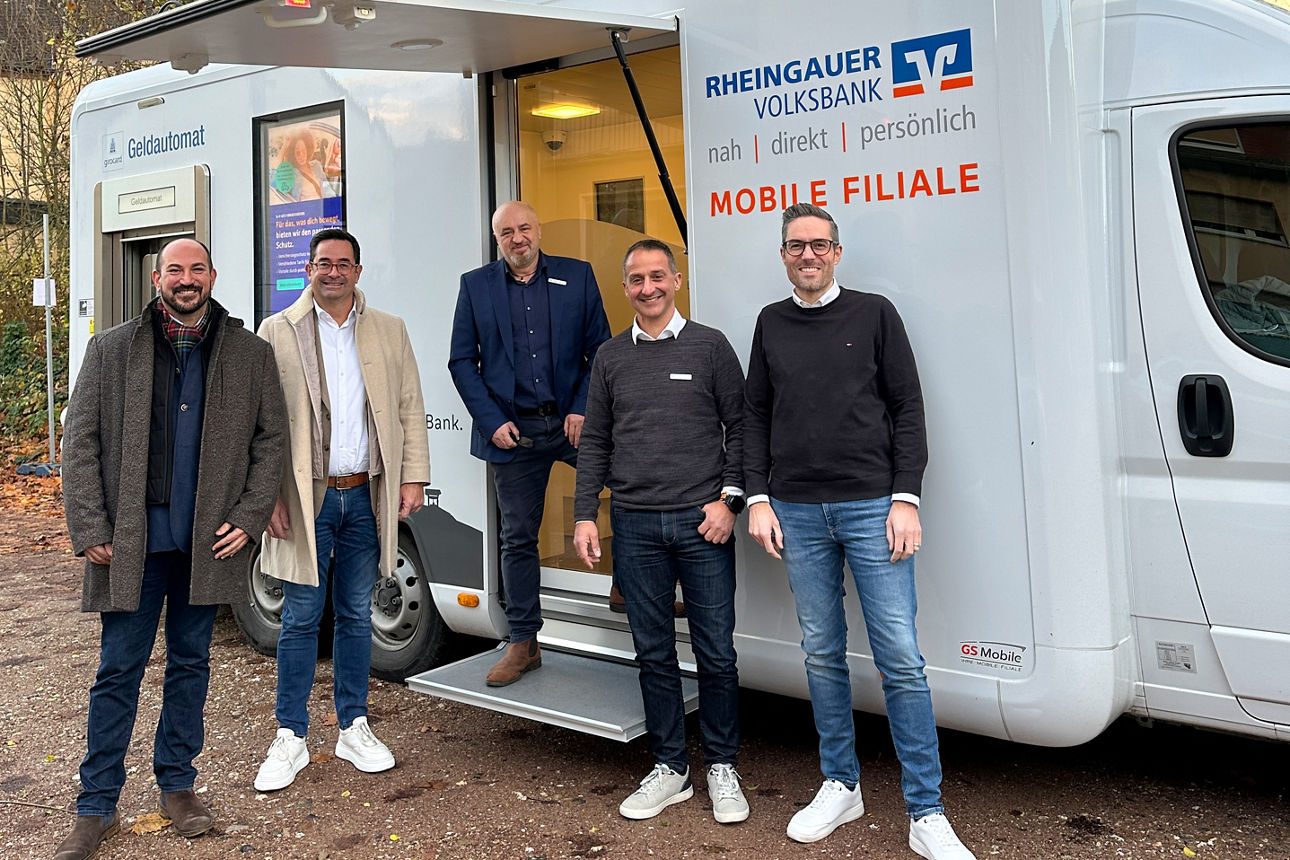 Fünf Männer stehen vor einem weißen mobilen Bankfahrzeug der Rheingauer Volksbank mit geöffnetem Seiteneingang.
