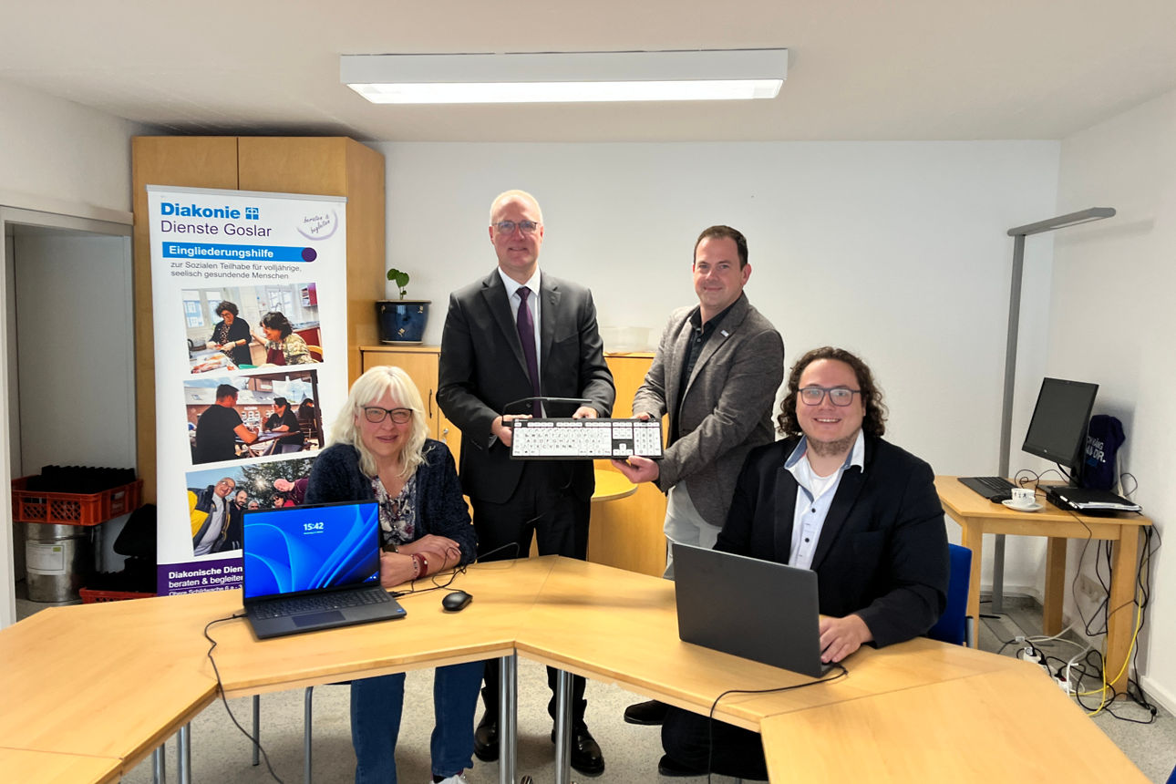 Vier Personen stehen oder sitzen um einen Holztisch. Zwei Männer in Anzügen stehen in der Mitte und halten gemeinsam eine Tastatur hoch. Die Dame links im Bild und der Herr rechts im Bild sitzen vor aufgeklappten Laptops. Im Hintergrund steht einer Banner der Diakonie Dienste Goslar zum Thema Eingliederungshilfe zur Sozialen  Teilhabe für volljährige, seelisch gesundende Menschen.