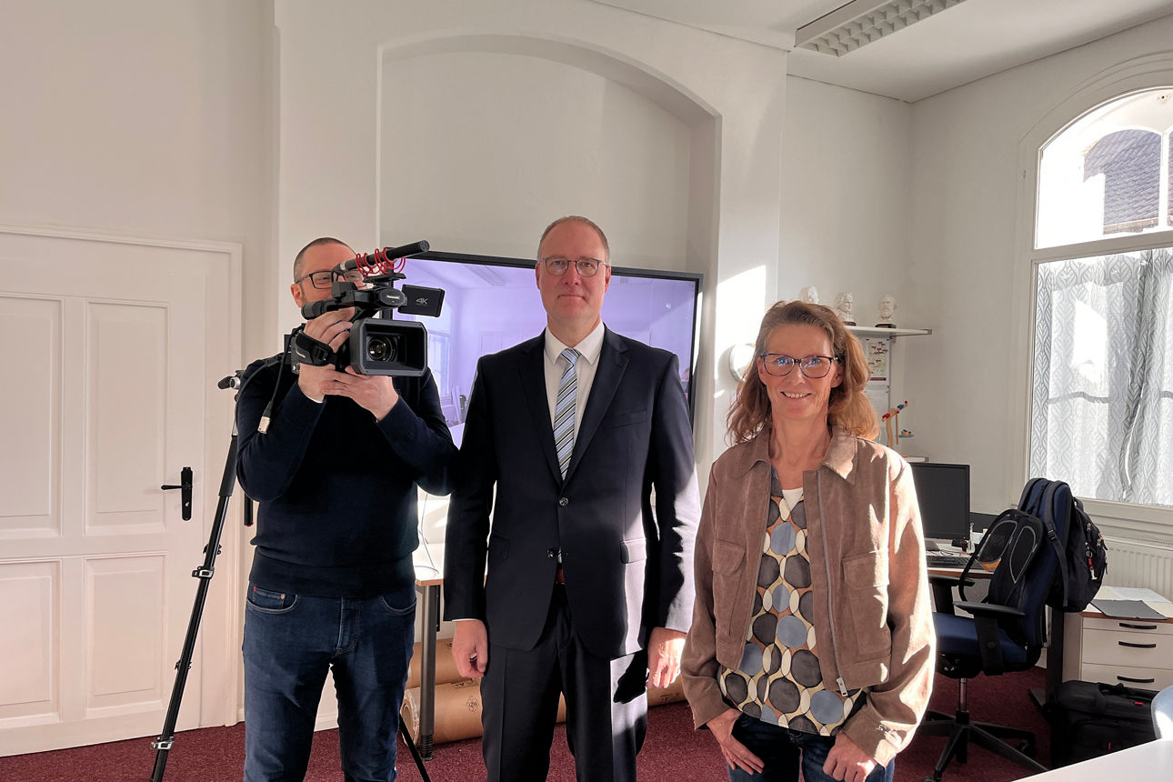Drei Personen, ein Kameramann, ein Mann im Anzug und eine Frau, posieren für eine Aufnahme vor einem großem Bildschirm. 