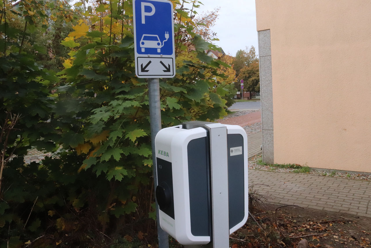 Ladesäule mit blauem Parkplatzschild im Hintegrund vor einem grünen Strauch