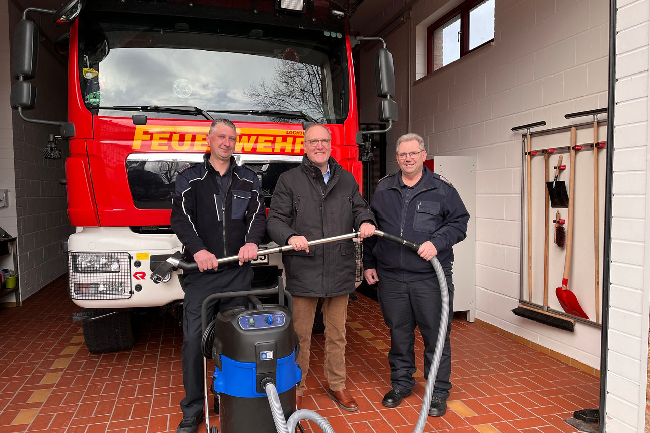 Drei Personen stehen vor einem roten Feuerwehrfahrzeug in einer Fahrzeughalle und präsentieren einen großen Industriesauger mit langem Schlauch. An der Wand hängen verschiedene Werkzeuge wie Besen und Schaufeln.