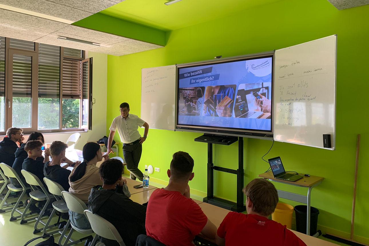 Ein Mann steht vor einer Schulklasse. Im Hintergrund ist eine Tafel zu sehen.