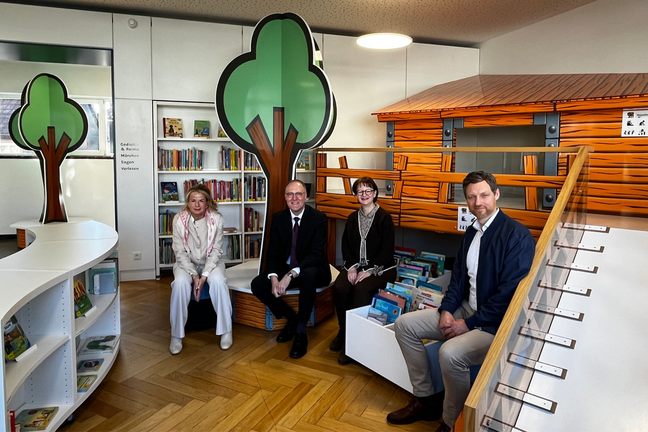 Freundlich gestaltete Kinderecke in einer Bibliothek. Vier Erwachsene auf einer niedrigen Bank vor Bücherregalen. Im Hintergrund ist sind dekorative Baumaufsteller und ein holzverkleidetes Spiel- oder Lesehaus mit einer kleinen Treppe auf der rechten Seite. Es ist ein toller heller Raum.