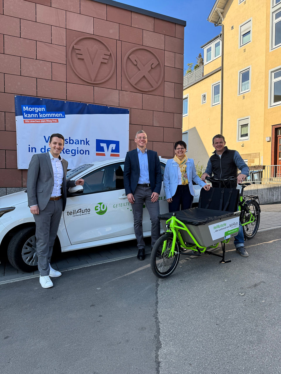 Drei Männer und eine Frau stehen mit Fahrrad und Auto vor dem Volksbank Gebäude