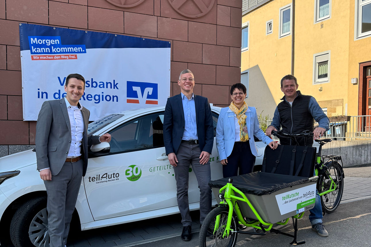 Drei Männer und eine Frau stehen mit Fahrrad und Auto vor dem Volksbank Gebäude