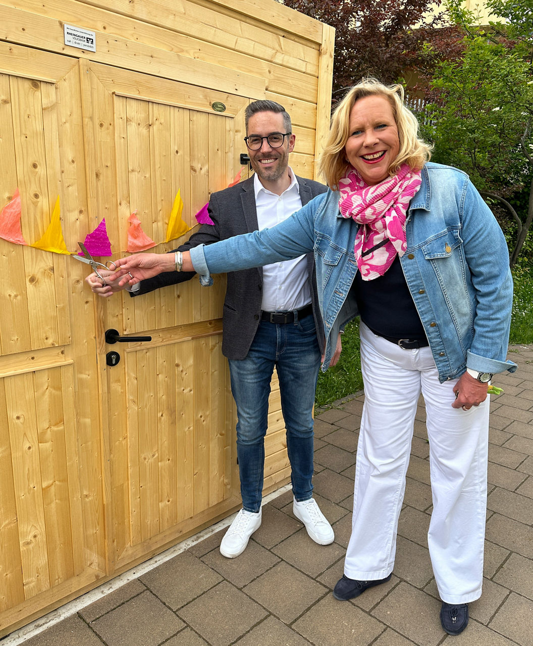 Zwei Personen vor einer Holztür, die ein Band mit bunten Papierfahnen zerschneiden, beide lässig gekleidet