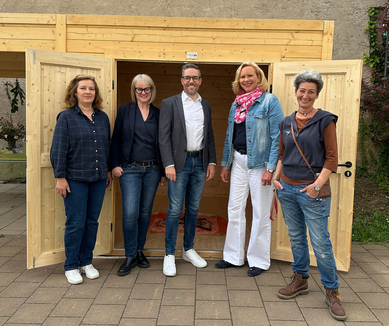 Fünf Personen stehen vor einer offenen Holztür einer neuen Gartenhütte aus Holz, alle tragen legere Kleidung und Schuhe.