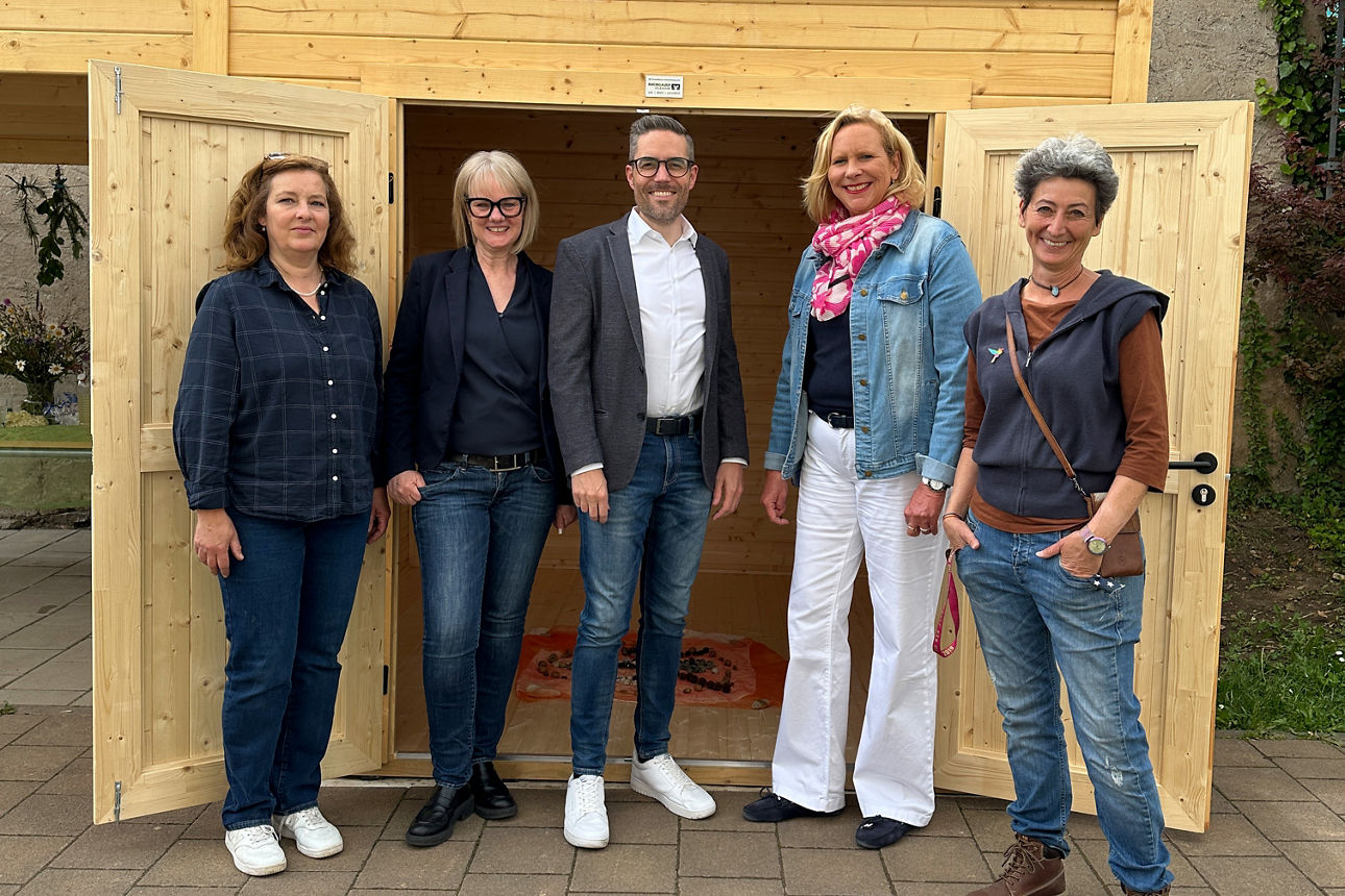 Fünf Personen stehen vor einer offenen Holztür einer neuen Gartenhütte aus Holz, alle tragen legere Kleidung und Schuhe.