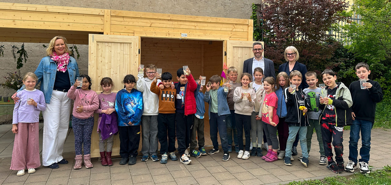 Gruppe von Kindern und drei Erwachsenen steht vor einer Gartenhütte aus Holz im Freien. Die Kinder halten Becher in der Hand