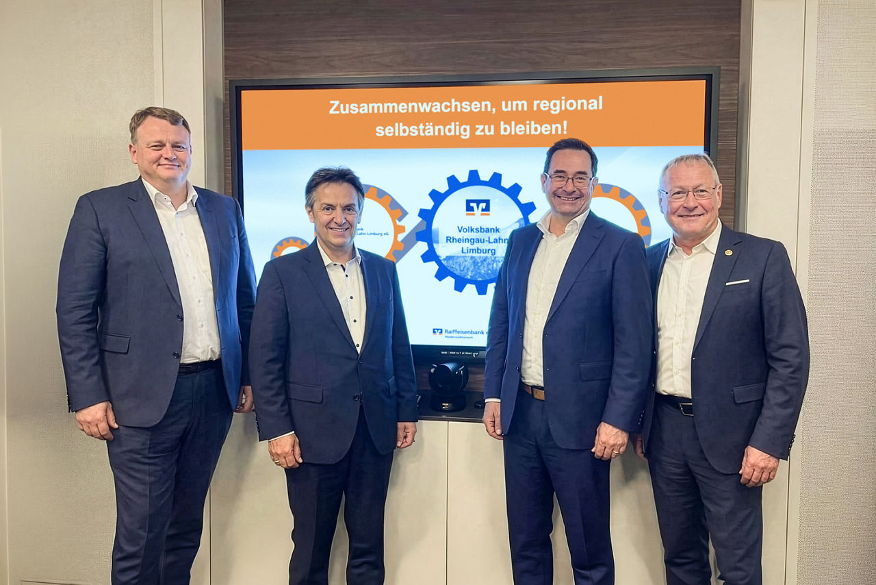 Vier Männer in Anzügen stehen vor einem Bildschirm mit dem Text 'Zusammenwachsen, um regional selbstständig zu bleiben!' und einem Logo der Volksbank Rheingau-Lahn-Limburg