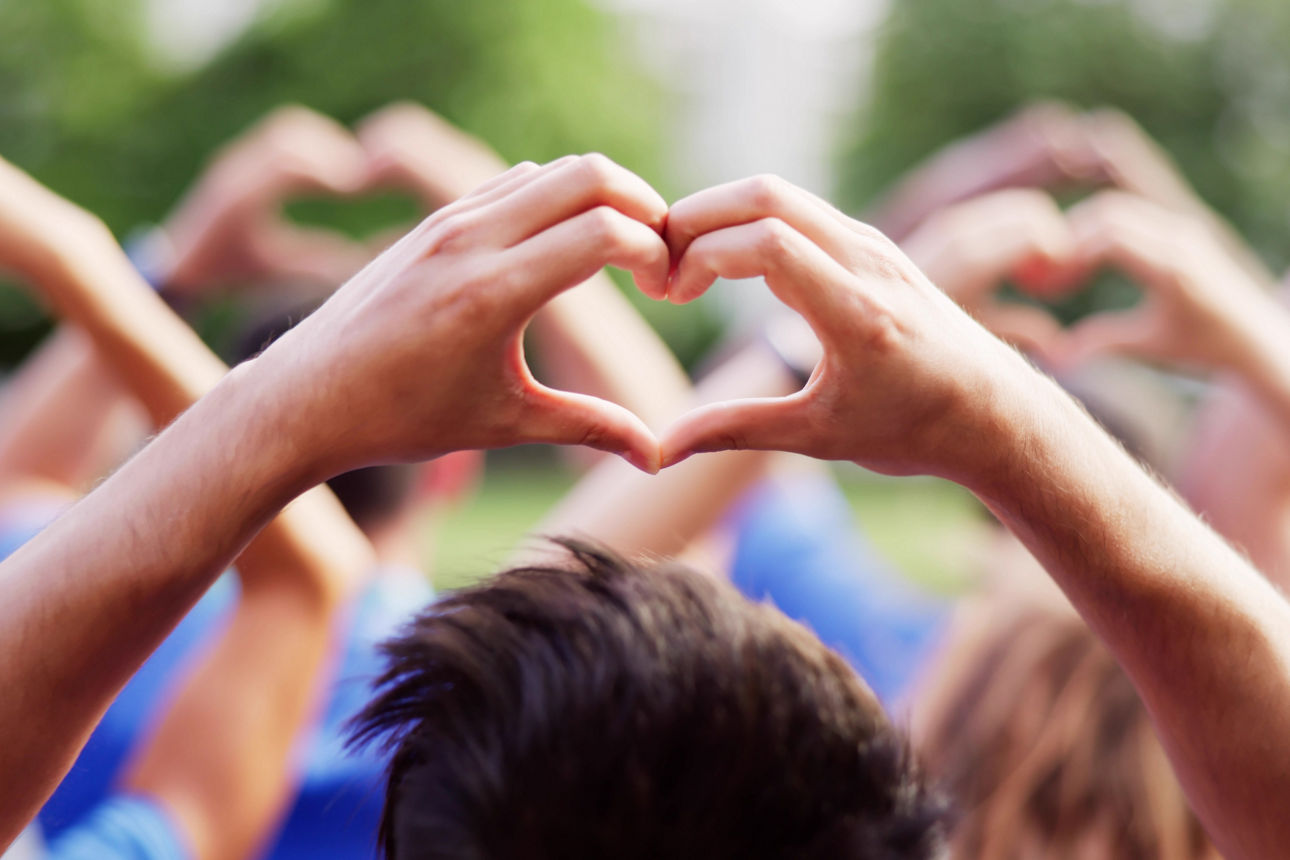 Mehrere Menschen formen mit ihren Händen Herz-Symbole, im Hintergrund sind weitere Personen unscharf zu erkennen.