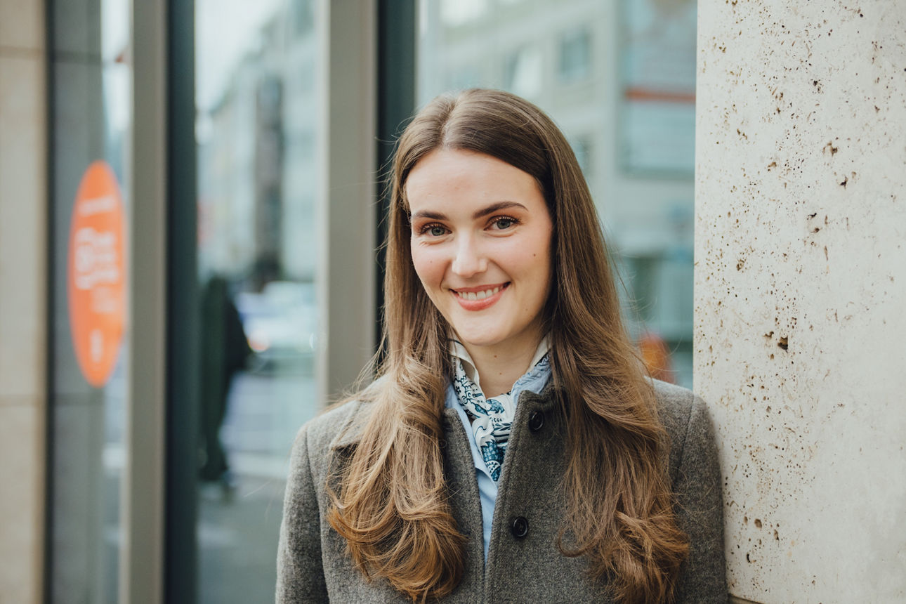 Eine Mitarbeiterin steht vor der Kölner FIliale und lacht in die Kamera