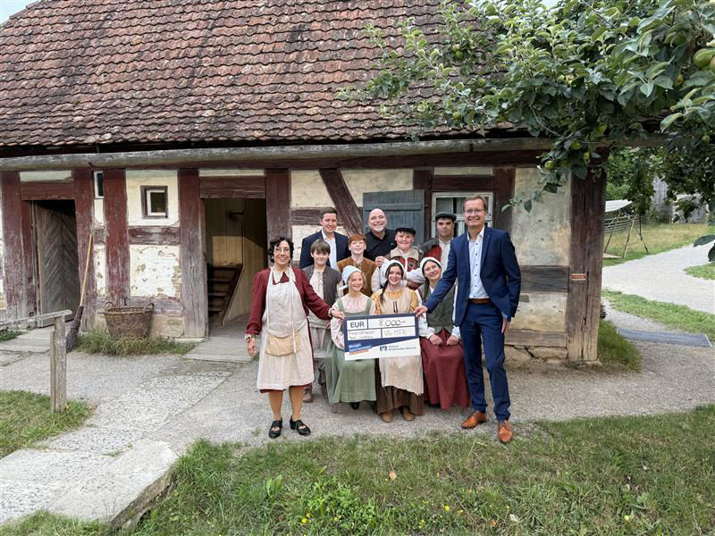 Eine Gruppe von Menschen in historischer Kleidung steht vor einem alten Fachwerkhaus und halten einen Scheck.