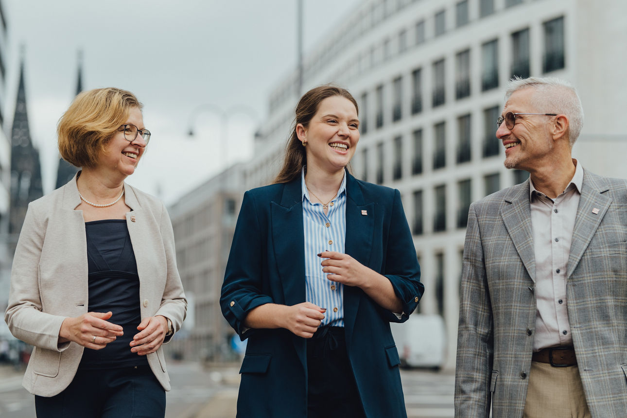 Drei Mitarbeitende der Pax-BKC unterhalten sich, im Hintergrund sieht man den Kölner Dom