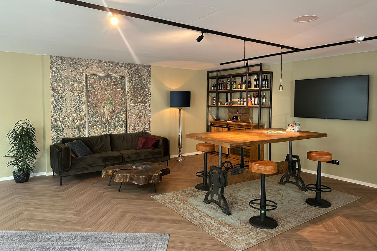 Zu sehen ist die stilvolle Innenansicht der Volksbank Immobilien in Gladbeck. Das Blick fällt auf den gemütlichen Wartebereich. Vor einer hellgrünen Wand, die als Highlight eine geschmackvolle Motivtapete aufweist, steht eine dunkelgrüne Couch, davor ein rustikaler Tisch, der aus einer Baumscheibe besteht. An der rechten Wand steht ein Regal mit Spetialitäten aus Mallorco, die zudem zum Verkauf angeboten werden. Neben dem Regal hängt ein großer Fenrseher an der Wand, davor steht ein rustikaler Tisch mit 4 Hockern im Industrie-Chic. 