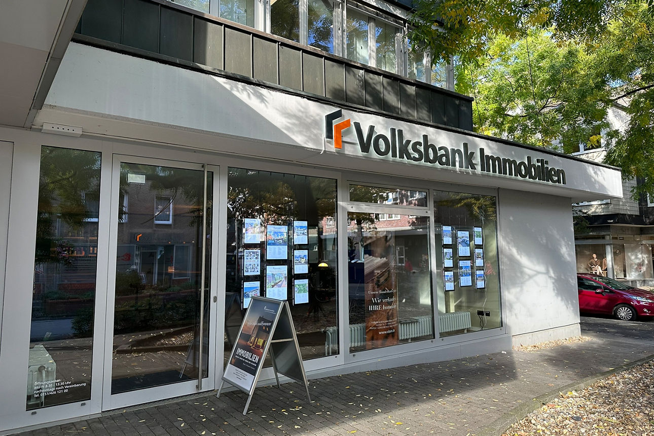 Zu sehen ist die Außenansicht der Volksbank Immobilien in Gladbeck. Sie zeichnet sich durch eine großzügige Glasfront mit bodentiefen Fenstern aus. Die Eingangstür ist ebenfalls aus Glas. In den Schaufenstern hängen mehrere beleuchtete Bilderrahmen, in denen unterschiedliche Immobilienexposés zu sehen sind. Vor der Tür steht ein A1-Aufsteller mit Plakaten. Der Eingang ist über einen barrierefreien Zugang erreichbar, der von der Lambertistraße abgeht.