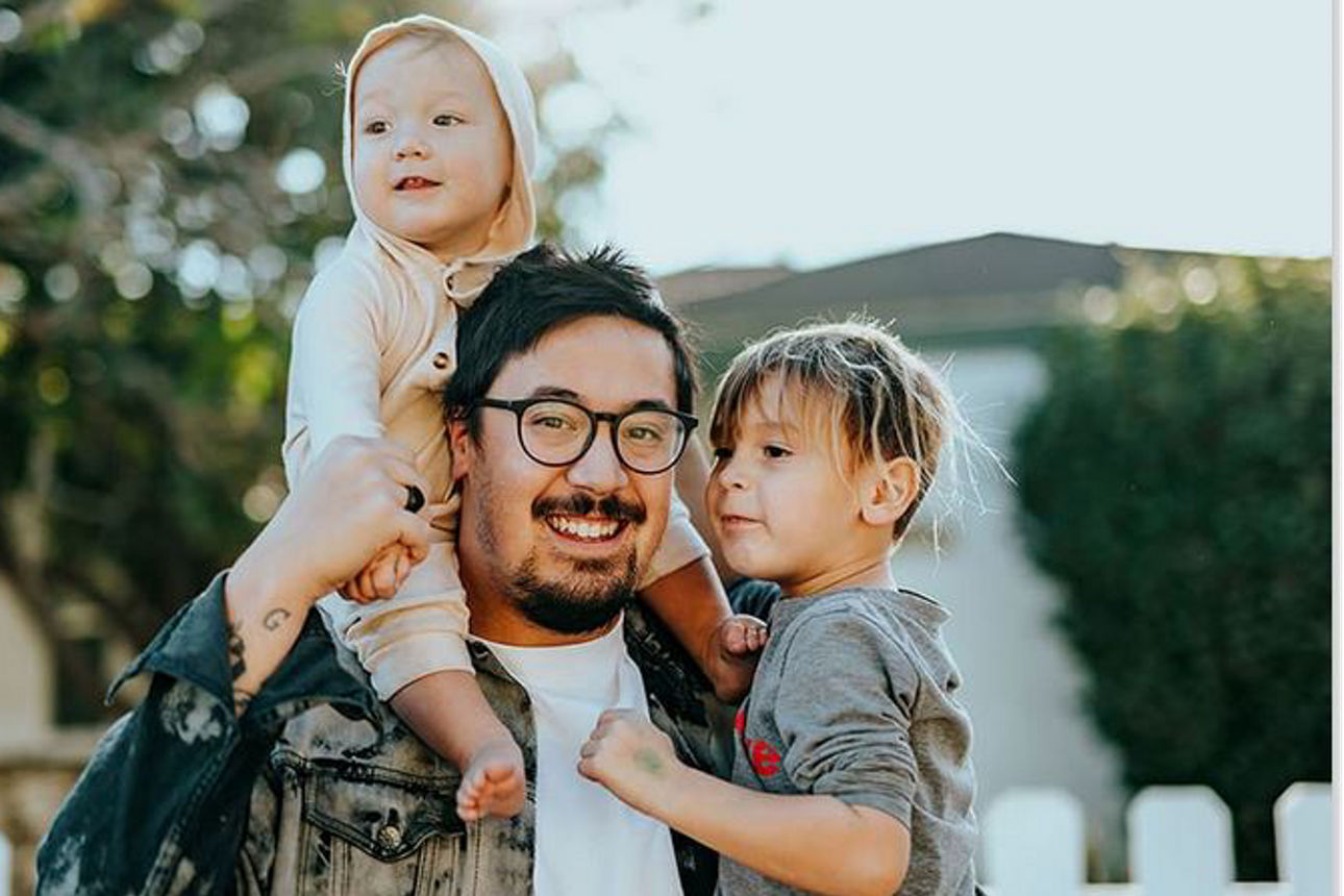 Ein Vater steht vor einem Gartenzaun mit zwei Kindern im Arm