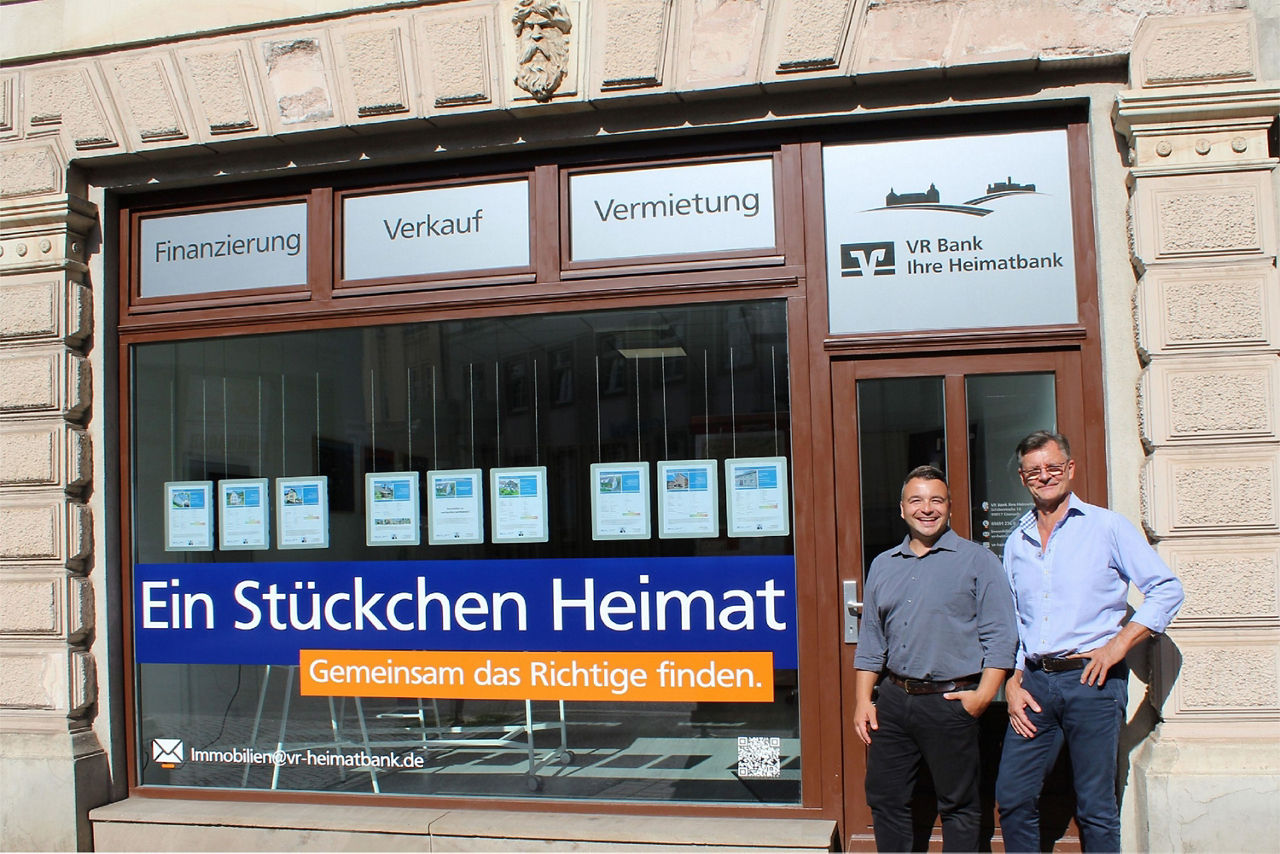 Zwei Männer stehen vor einem Gebäude der VR Bank Ihre Heimatbank im Schaufenster mehrere Immobilienangebote