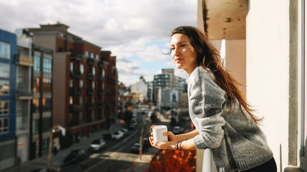Eine junge Frau steht mit einer Kaffeetasse auf einem Balkon und blickt der Sonne entgegen