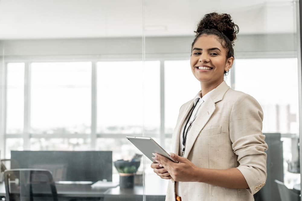 Eine junge Frau im Business-Outfit hält ein Tablet in der Hand und lächelt in die Kamera