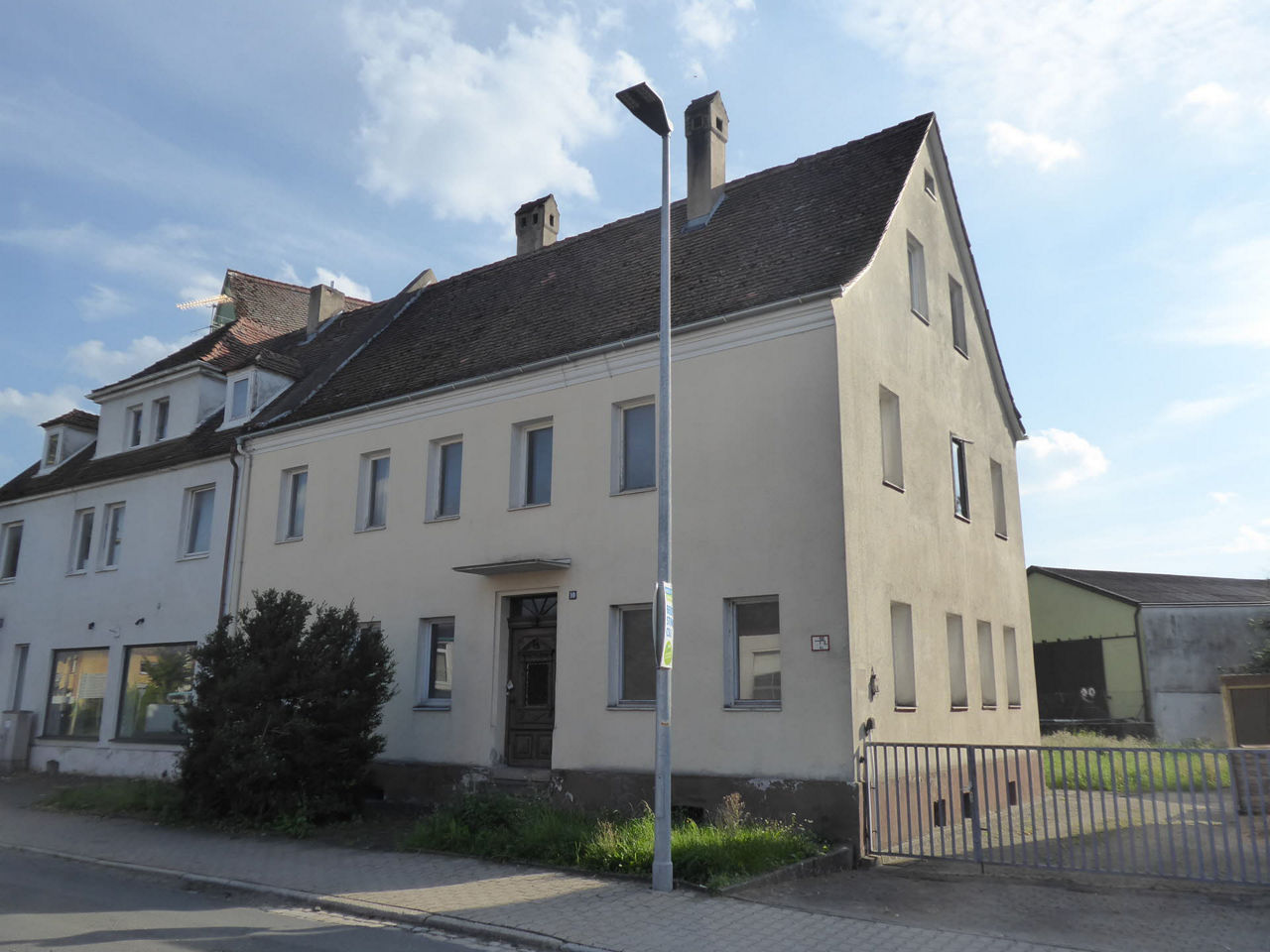 Bild von Häuserfront, im Vordergrund sieht man einen Busch, eine Straßenlaterne und den Gehweg.