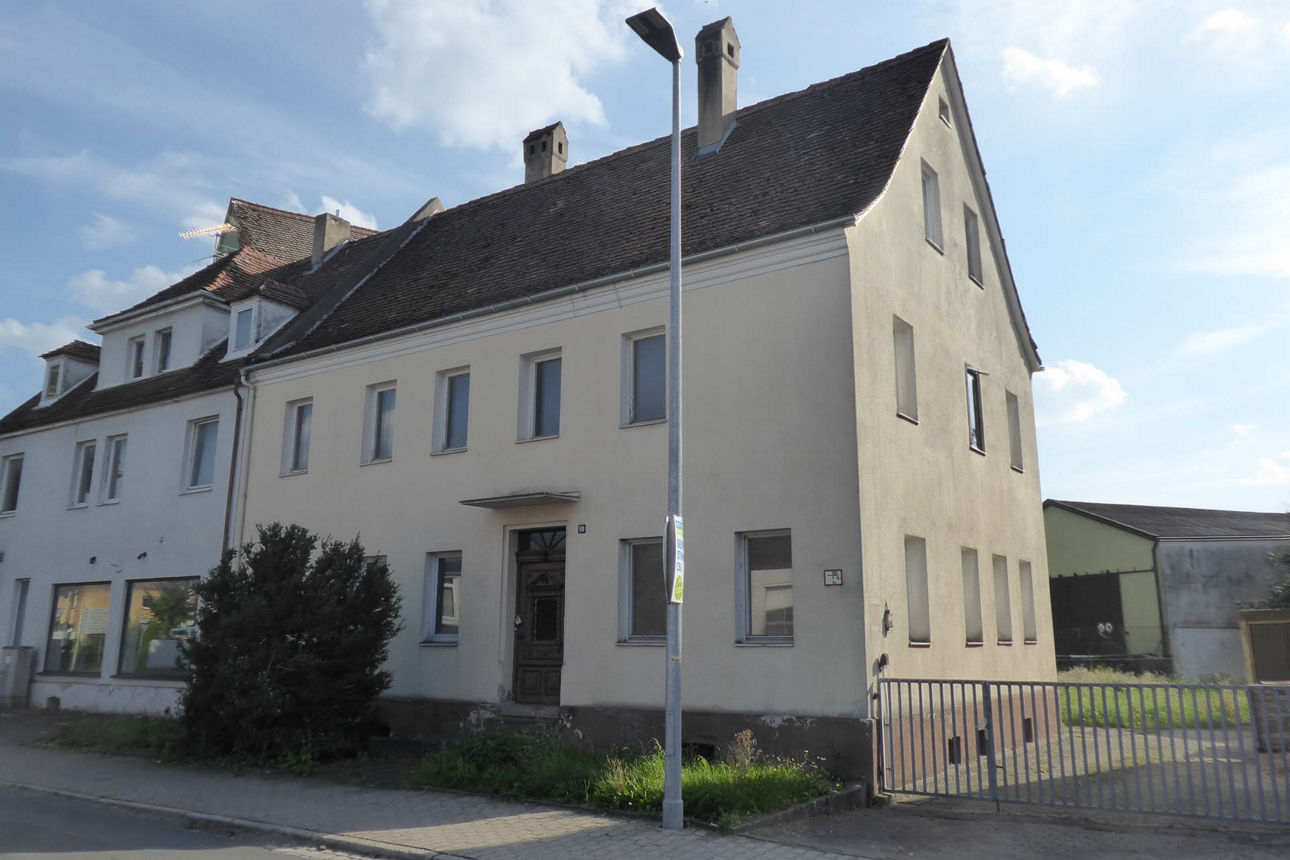 Bild von Häuserfront, im Vordergrund sieht man einen Busch, eine Straßenlaterne und den Gehweg.