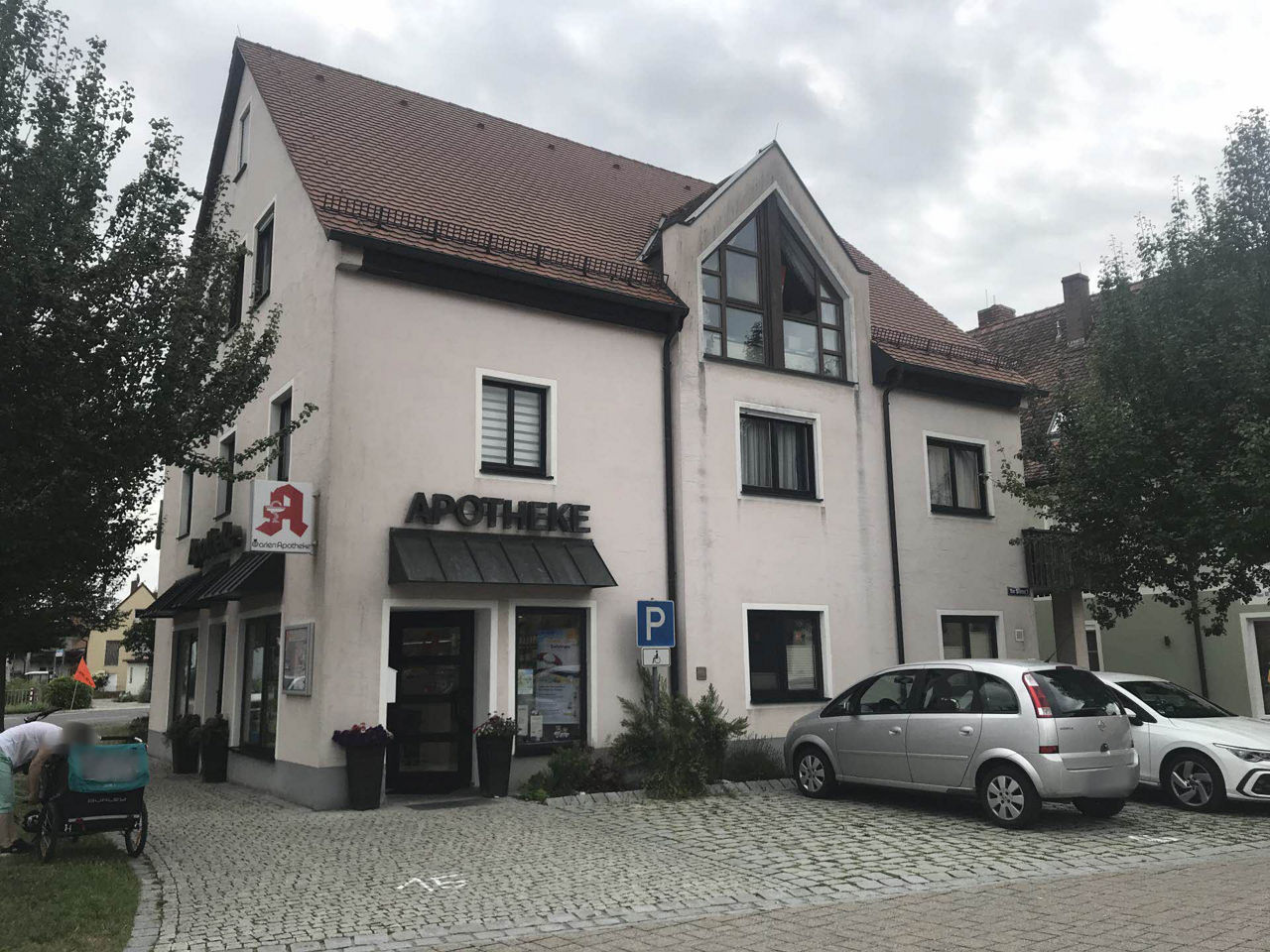 Bild von dreistöckigem Haus, eingerahmt von Bäumen. Im Vordergrund auf gepflasterten Parkplätzen parkten zwei PKW