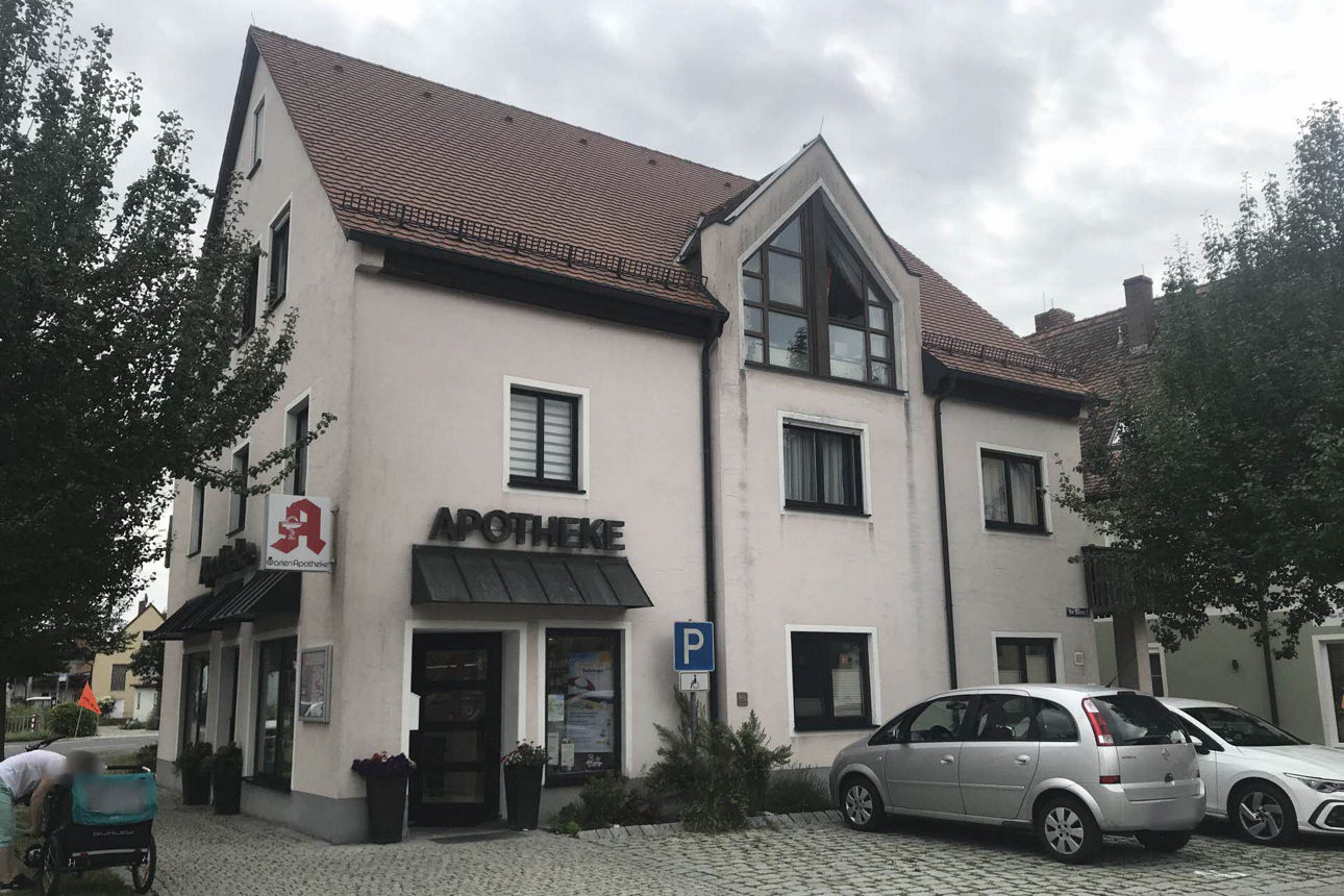 Bild von dreistöckigem Haus, eingerahmt von Bäumen. Im Vordergrund auf gepflasterten Parkplätzen parkten zwei PKW