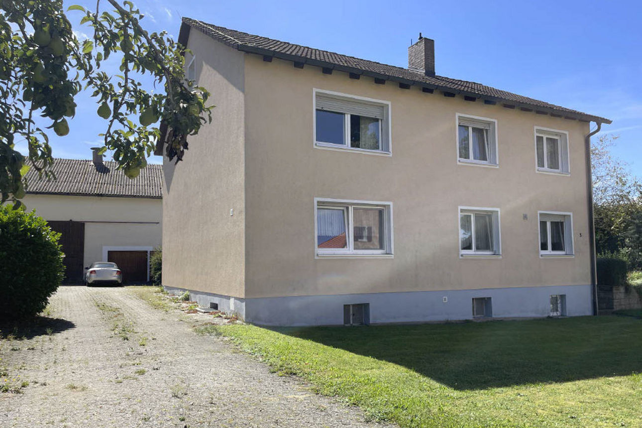 Ein zweistöckiges, beiges Wohnhaus mit mehreren Fenstern und einer gepflasterten Hofeinfahrt