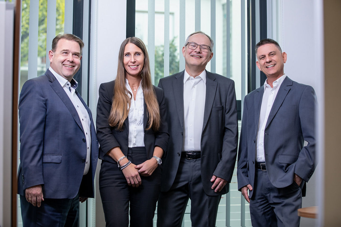 Eine Frau und drei Männer in Anzügen lächeln in die Kamera