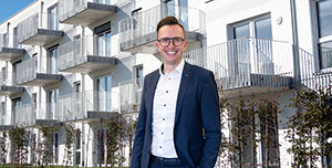 Immobilienvermittler Simon Weiß vor einem großen weißen Gebäude. Er trägt einen Anzug.