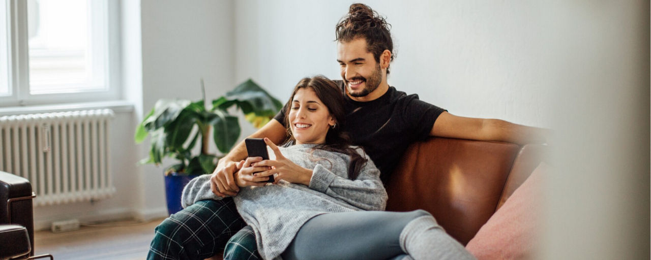 Junges Pärchen liegt auf der Couch und schaut in ein Smartphone
