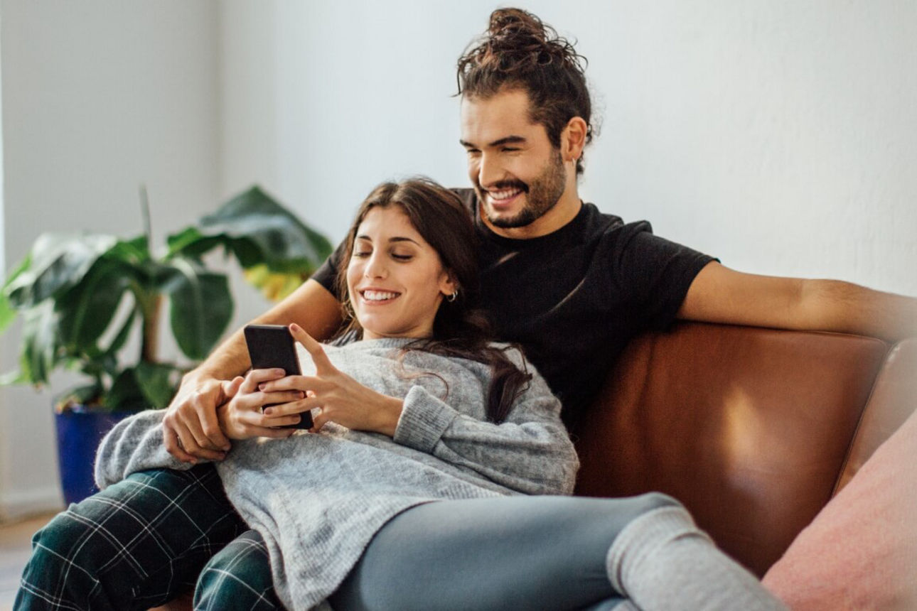 Junges Pärchen liegt auf der Couch und schaut in ein Smartphone
