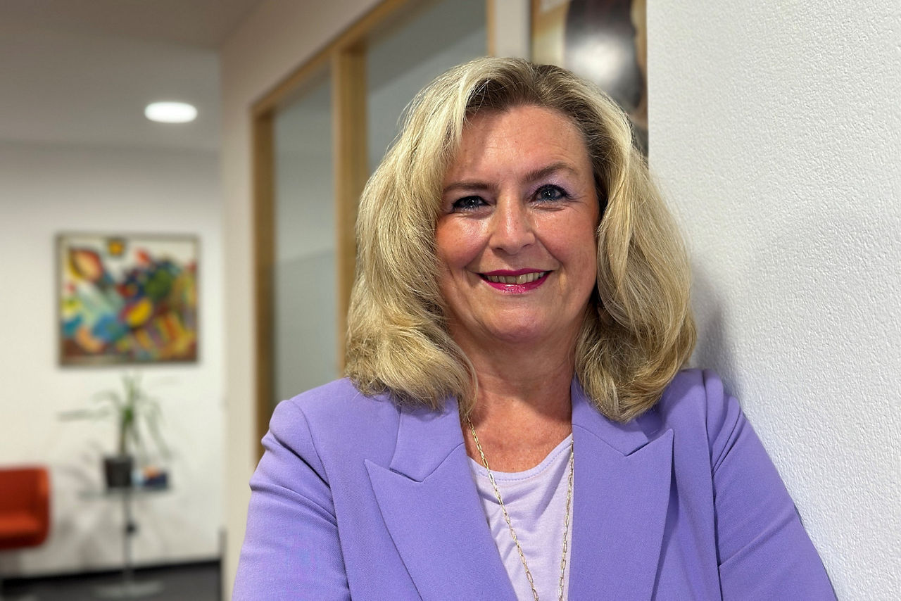 Eine blonde Frau in einem lila Blazer steht lächelnd vor einer weißen Wand in einem modern eingerichteten Büro.