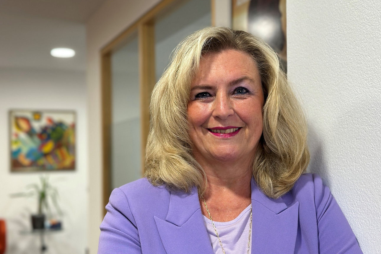 Eine blonde Frau in einem lila Blazer steht lächelnd vor einer weißen Wand in einem modern eingerichteten Büro.