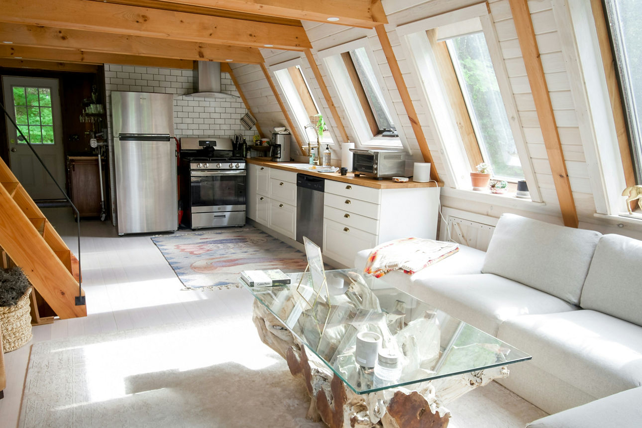 Blick in eine Wohnung mit Holztreppe und Holzbalken, helle Couch und Küche im Hintergrund.