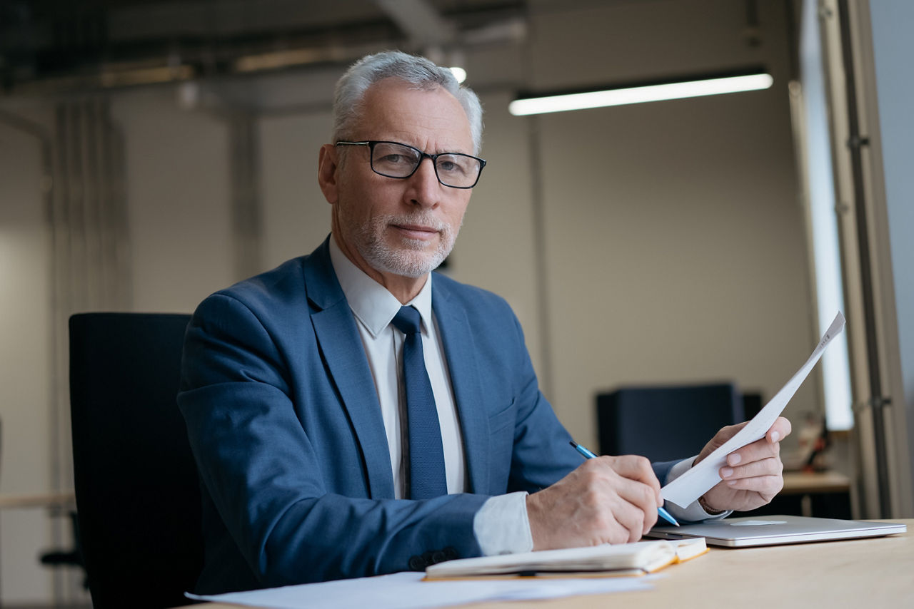 Geschäftsmann mit Papierdokumenten in einem modernen Büro. Porträt eines nachdenklichen Senior-Managers in formeller Kleidung, der am Arbeitsplatz in die Kamera blickt. 