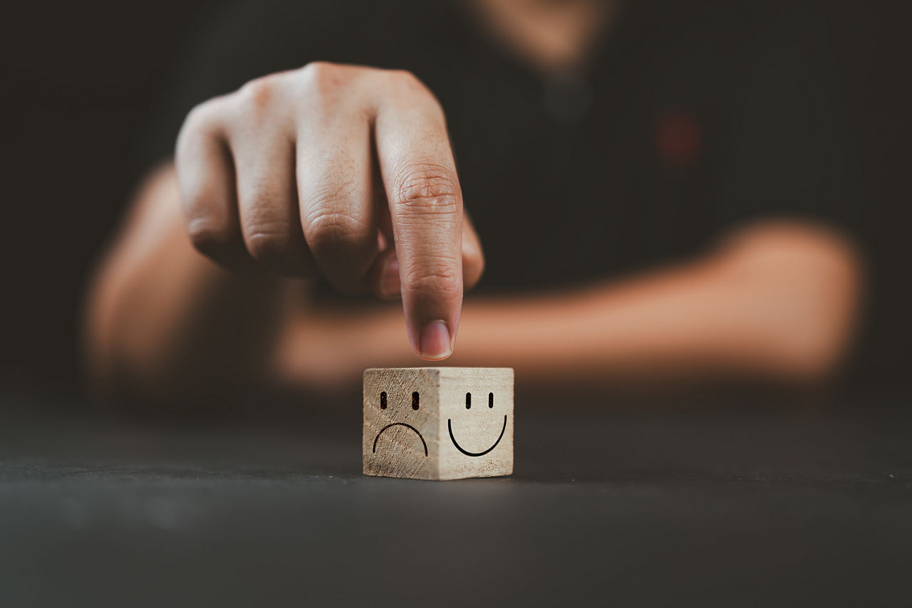 Die Hand eines Mannes zeigt auf einen Holzwürfel mit einem glücklichen Lächeln auf der hellen Seite und einem unglücklichen Gesicht auf der dunklen Seite, um eine positive Einstellung zu erreichen.