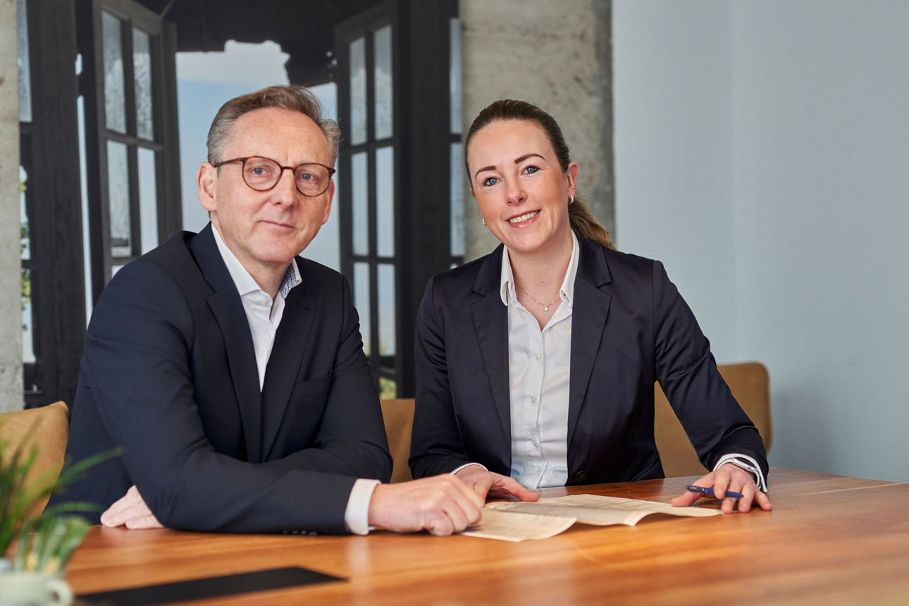 Mann und Frau sitzen nebeneinander an einem Tisch und blicken in die Kamera.