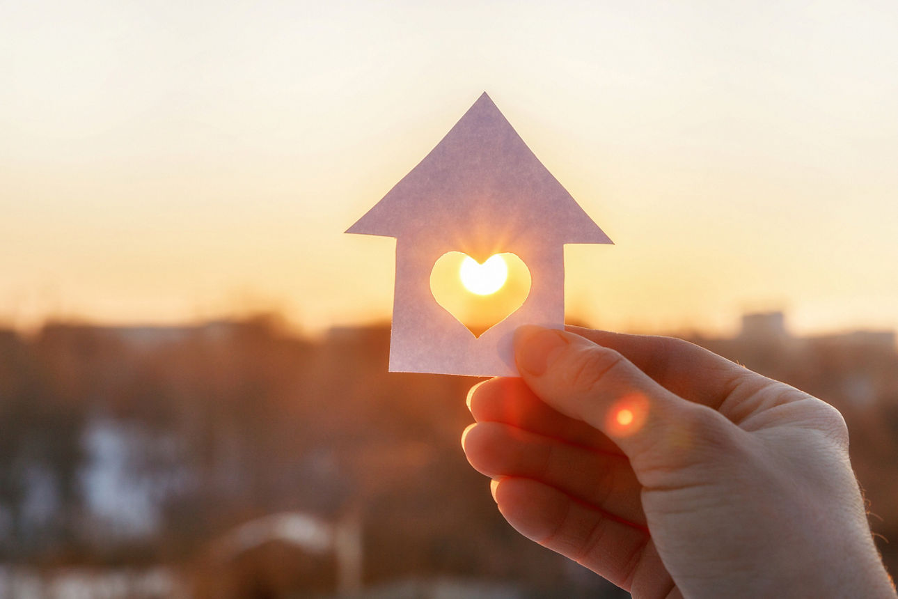 Eine Hand, welche ein Haus aus Papier mit einem Herz in der Mitte festhält. 