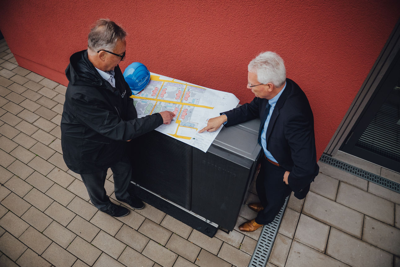 Ein Mitarbeiter und ein Kunde stehen vor einem Gebäude und schauen sich einen Bauplan an