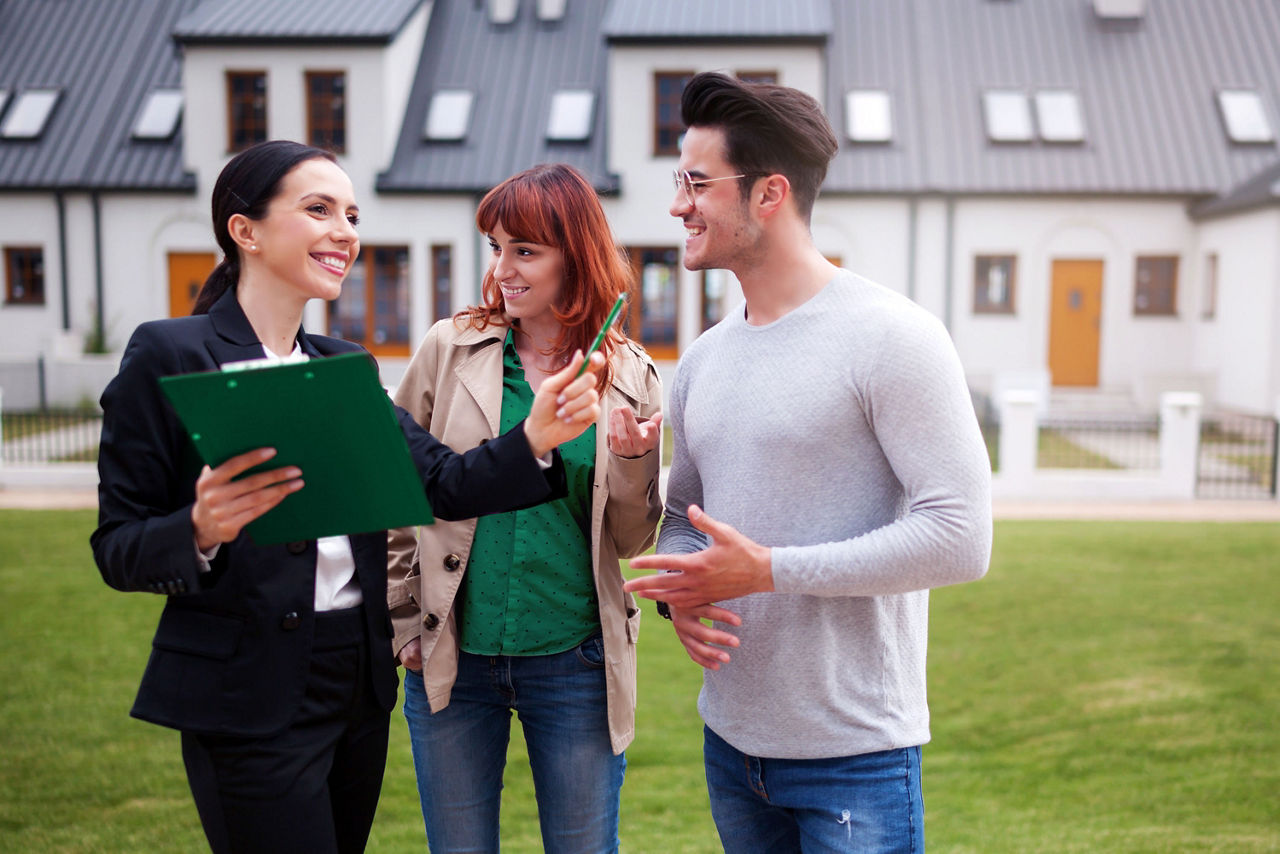 Eine Immobilienmaklerin steht mit einem Paar vor einer Häuserreihe und zeigt auf ein Haus.