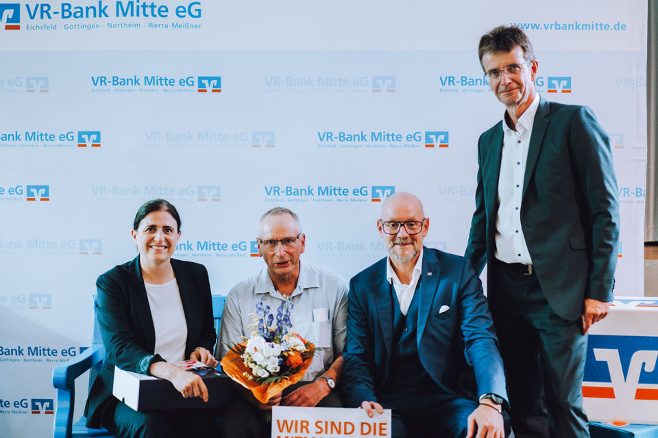Drei Menschen sitzenauf einer blauen Bank, eine Person steht daneben.