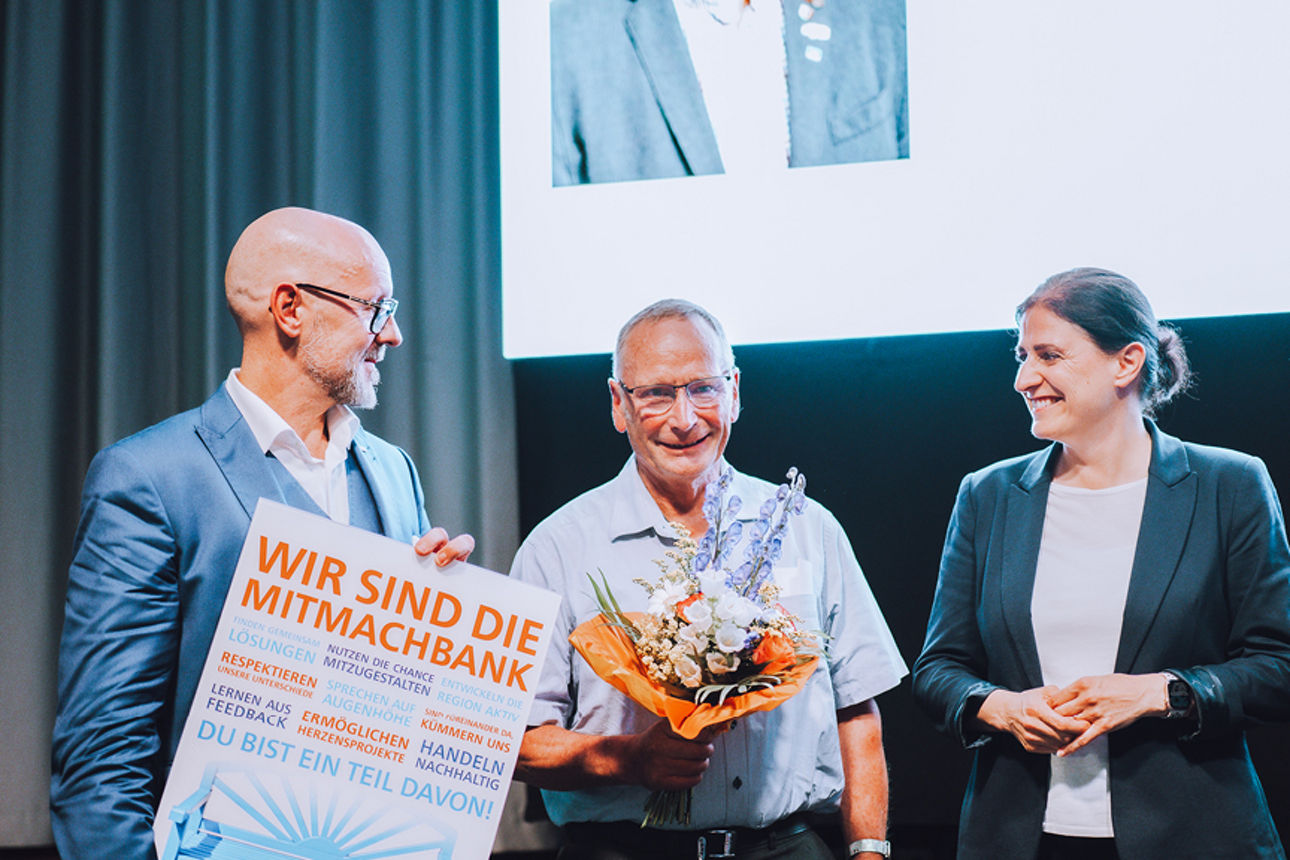 Drei Personen auf einer Bühne, einer mit blumenstrauß und iener mit einem Bild in der Hand
