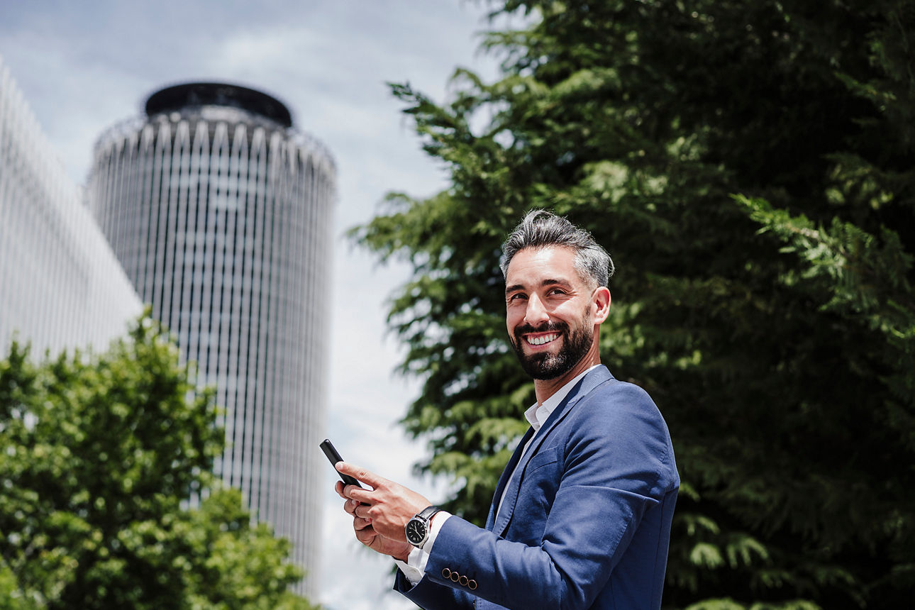 Smiling businessman with mobile phone standing in city on sunny day