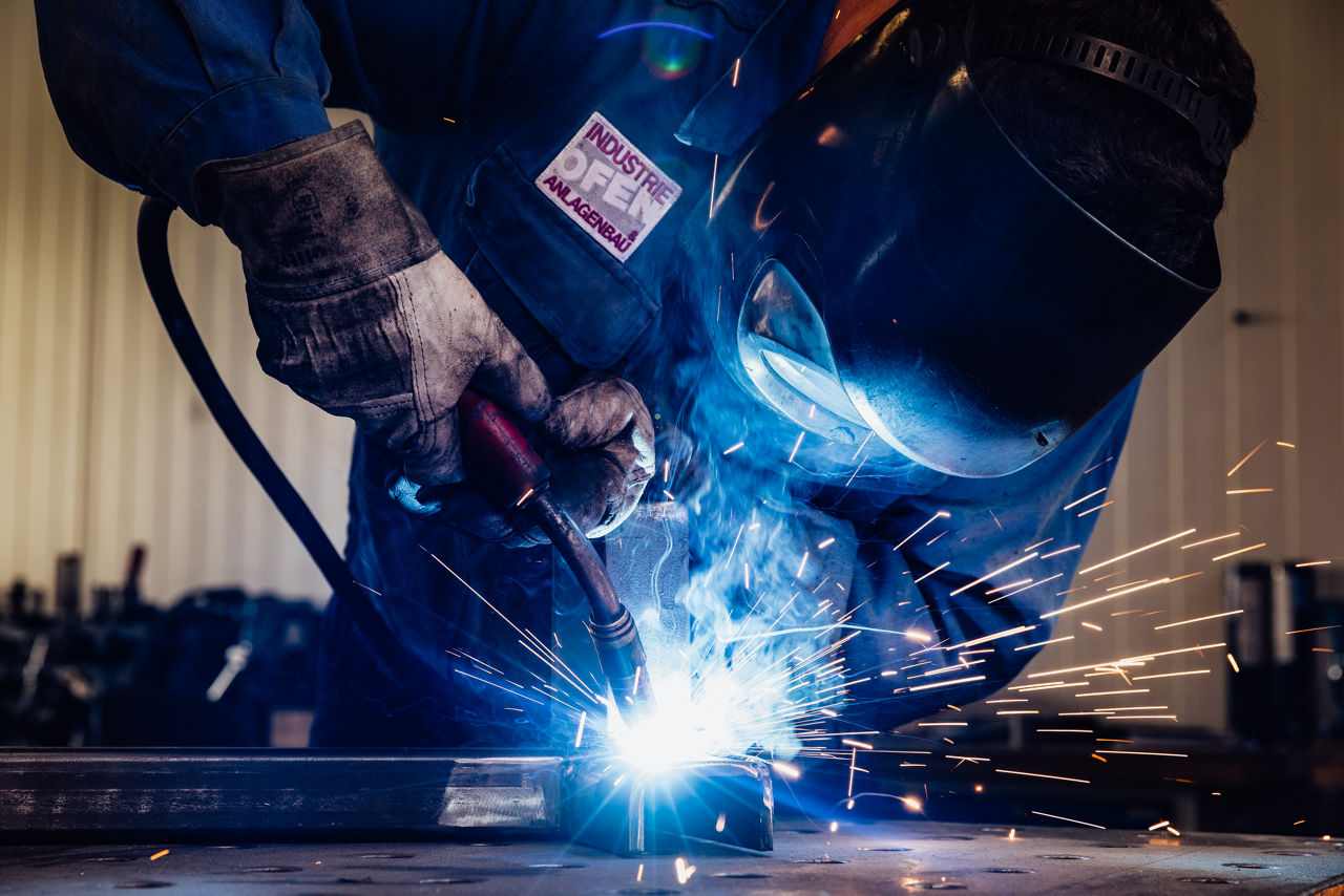 Ein Schweißer mit Schutzmaske und Schutzhandschuhen arbeitet an einem Metallstück, Funken sprühen beim Schweißen.