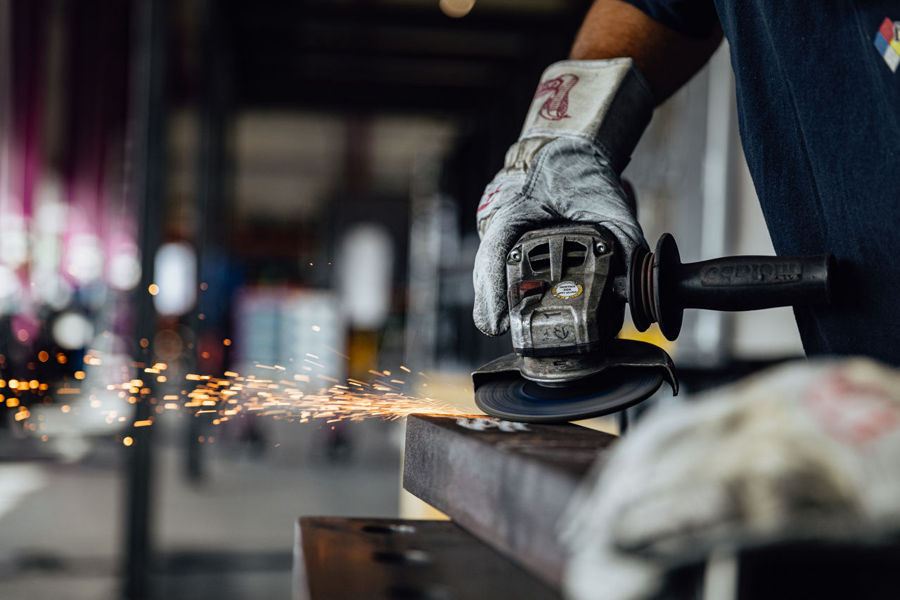 Ein Arbeiter mit Schutzhandschuhen schleift eine Metallkante mit einem Winkelschleifer, Funken sprühen beim Schleifen.