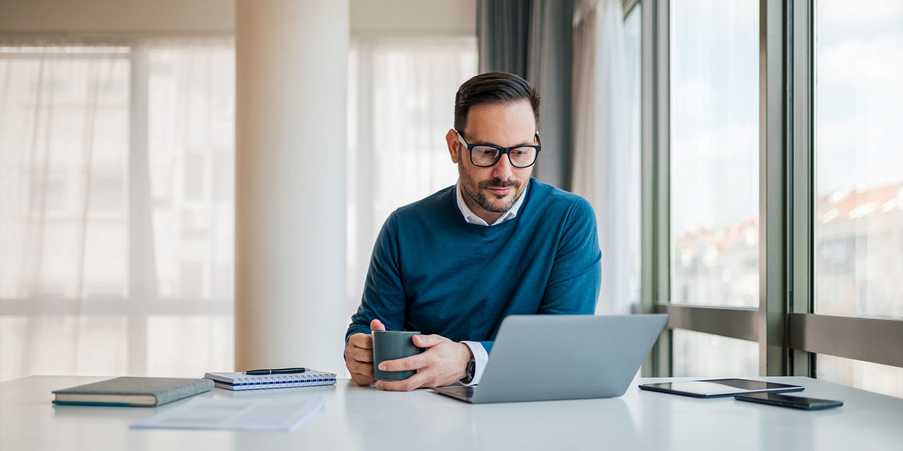 Ein Mann mit Brille sitzt konzentriert am Laptop und trinkt Kaffee